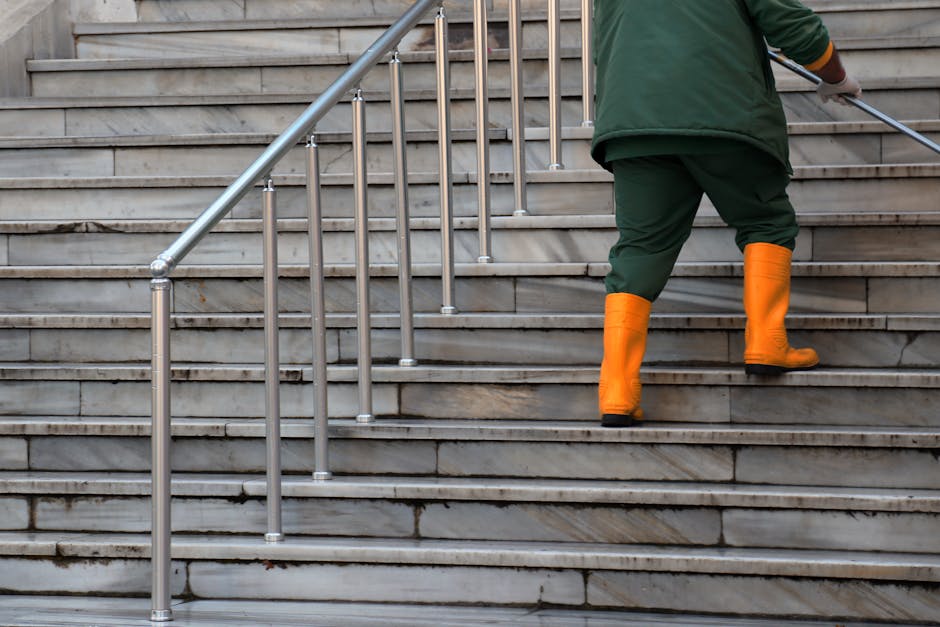 A person wearing green overalls and bright orange waterproof boots is ascending outdoor stone stairs with a metal handrail, which appears to be part of a building entrance. The stairs are made of grey