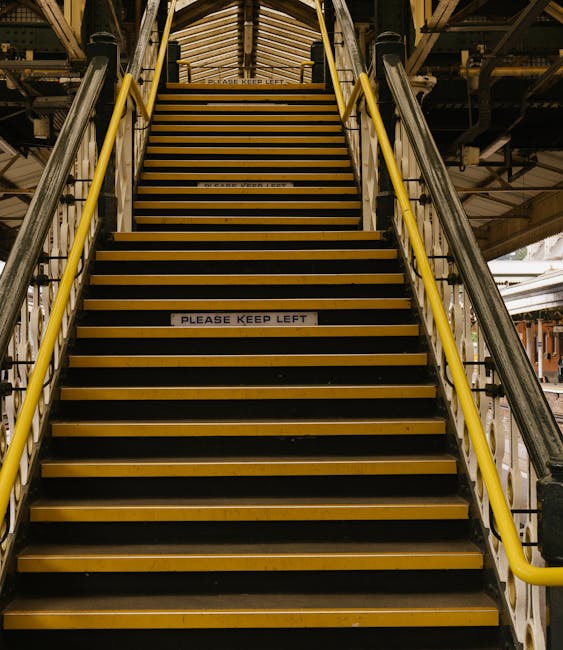 A person wearing green overalls and bright orange waterproof boots is ascending outdoor stone stairs with a metal handrail, which appears to be part of a building entrance. The stairs are made of grey