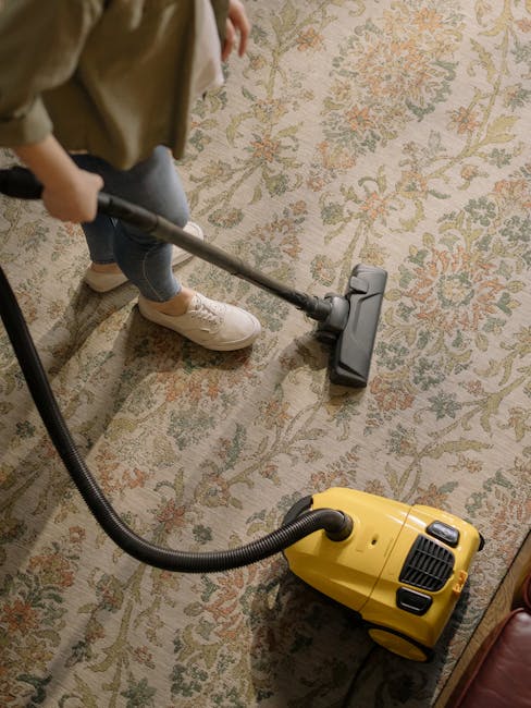A person wearing beige shoes and dark jeans is vacuuming a patterned, beige and green carpet in a residential room. The carpet appears clean and well-maintained, with a floral design visible. The vacuum cleaner, with a yellow body and black hose, is positioned on the carpet, suggesting an ongoing deep cleaning process. Natural lighting illuminates the area, highlighting the surface's tidy state. This cleaning setup exemplifies domestic surface cleaning, emphasizing hygiene and maintenance. The scene is part of a routine cleaning session by Cleaners Kingston upon Thames, demonstrating effective carpet and floor sanitisation.