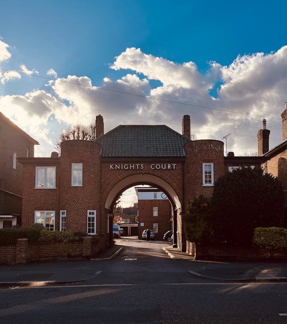 Photograph of the entrance to Knights Court, a residential building with a brick archway, located on Kingston High Street KT1. The structure features red brick walls, with the name 'Knights Court' displayed above the arch. The building has small, white-framed windows and is surrounded by greenery, including neatly trimmed bushes. The sky above is partly cloudy with patches of blue, and the scene is illuminated by natural daylight. This setting exemplifies a well-maintained, traditional residential area in Kingston upon Thames, with the potential for detailed surface cleaning and home hygiene enhancement by experts such as Cleaners Kingston upon Thames, to maintain cleanliness and hygienic standards in similar properties.