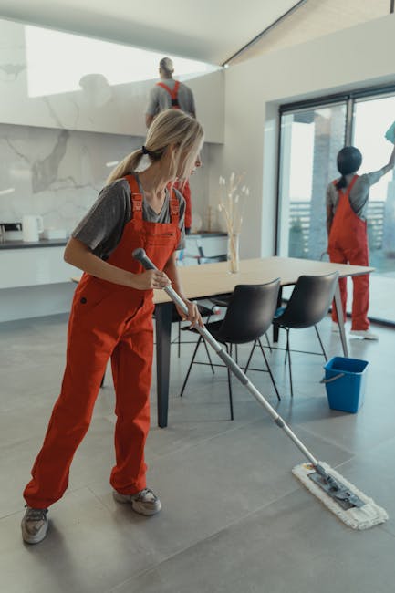 A female cleaner from Cleaners Kingston upon Thames is performing surface cleaning in a modern dining area with a mop, highlighting a deep cleaning process. The room features a large marble wall with natural veining, a glass sliding door revealing an urban view, and a polished grey tile floor that appears recently mopped with a wet shine. The dining table, made of light wood, is accompanied by black plastic chairs, and a blue cleaning bucket is positioned nearby. Bright, natural light floods the space through the large windows, emphasizing the clean and hygienic condition of the surfaces. Two other cleaners in matching red overalls are visible in the background, indicating a team effort in maintaining high standards of sanitisation, as part of professional domestic cleaning services provided by Cleaners Kingston upon Thames. The image showcases the company's commitment to surface and deep cleaning to ensure hygienic, spotless environments in residential or commercial settings.