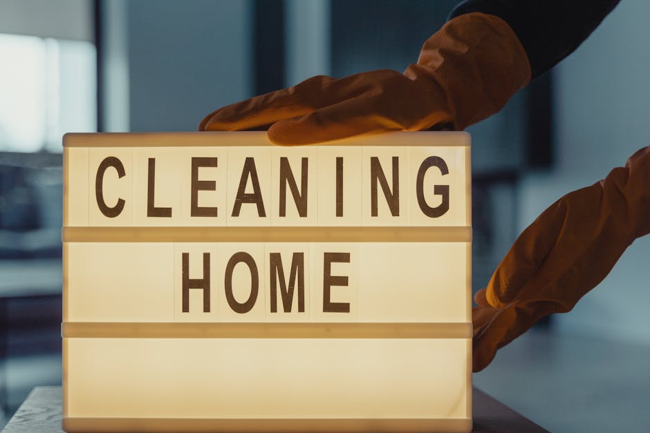 A person wearing orange cleaning gloves is placing or adjusting a lightbox sign that displays the words 'CLEANING HOME' in black capital letters on illuminated white background panels. The sign is situated on a surface in a room with blurred background elements, possibly a commercial or residential space. The scene emphasizes professional surface cleaning and sanitisation, aligning with services offered by Cleaners Kingston upon Thames, as referenced on the page for end of tenancy cleaning near Rose Theatre Kingston KT1, Kingston upon Thames. The lighting highlights the sign, creating a clean and organized atmosphere suitable for domestic cleaning or deep cleaning services.