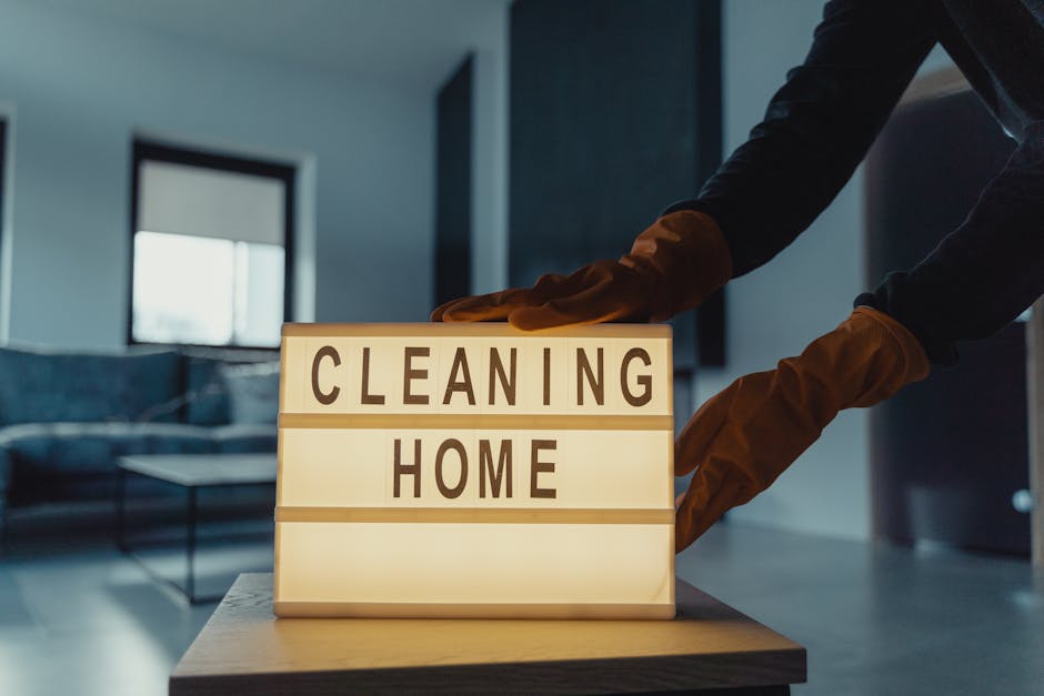 Close-up of a person wearing brown cleaning gloves, arranging a lightbox sign with the words 'CLEANING HOME' in a modern living room with a dark-tone couch, coffee table, and large window in the background. The scene is illuminated by natural daylight, highlighting the cleanliness and neatness of the space, which is part of a professional domestic cleaning service offered by Cleaners Kingston upon Thames, specializing in surface cleaning, deep cleaning, and sanitisation for residential properties near the Rose Theatre Kingston KT1 in Kingston upon Thames.
