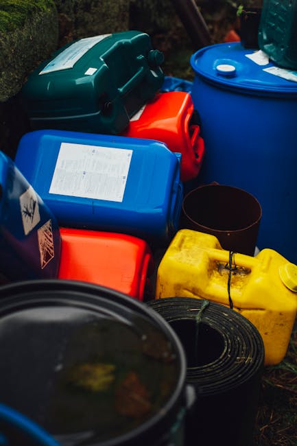 An outdoor scene featuring a collection of colourful hazardous waste containers, including blue, red, yellow, and black, placed on the ground with some stacked or leaning against each other. The conta