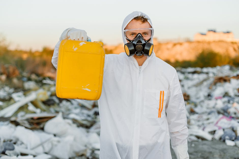An outdoor scene featuring a collection of colourful hazardous waste containers, including blue, red, yellow, and black, placed on the ground with some stacked or leaning against each other. The conta