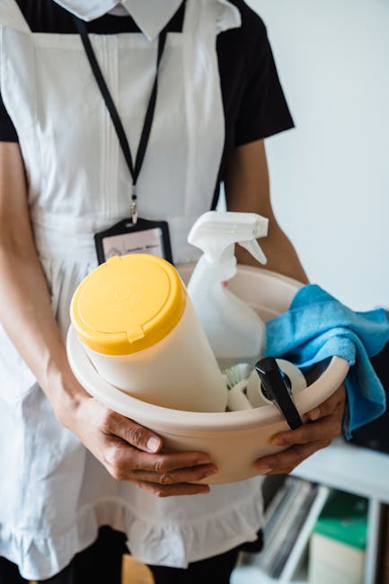 A person dressed in a black and white uniform holding a beige cleaning caddy containing various cleaning supplies, including a yellow spray bottle, a white spray bottle, a blue cleaning cloth, and a small black device, in an indoor setting. The background features a white wall and shelves with organized cleaning tools, reflecting a professional environment dedicated to surface cleaning, sanitisation, and domestic cleaning services offered by Cleaners Kingston upon Thames, aimed at maintaining hygiene and cleanliness for residential or commercial spaces.