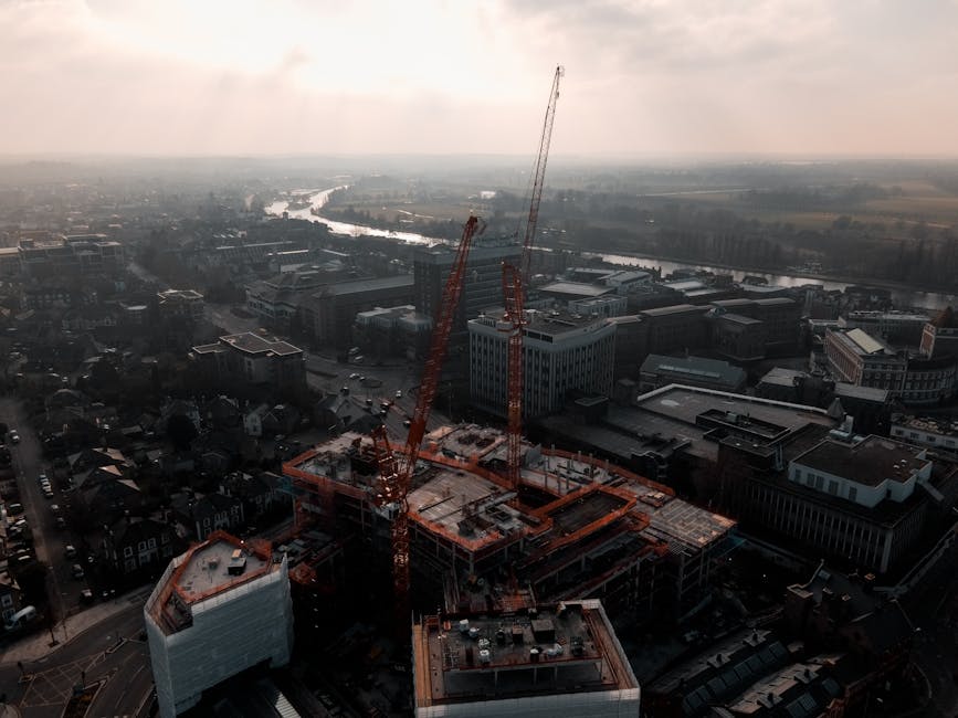 Aerial view of a construction site in an urban area with several cranes actively lifting materials, surrounded by modern glass and concrete buildings, under a cloudy sky during daytime. The site shows exposed surfaces of the building framework, with visible construction equipment, scaffolding, and materials. Adjacent streets feature parked cars and nearby residential and commercial structures. The scene highlights the ongoing development process within the cityscape, emphasizing a busy construction environment. Cleaners Kingston upon Thames emphasizes professional surface cleaning and deep cleaning services for commercial buildings such as these, ensuring proper sanitisation during or after construction projects.