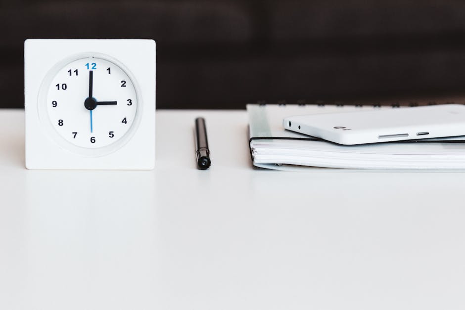 A close-up of a white square clock showing the time as 2:00, a black pen placed horizontally next to it, and a neatly stacked pile of white papers and a smartphone with a white case resting on top, all arranged on a clean, white surface. The background features a dark wooden surface, highlighting the organized and tidy appearance of a workspace. The image emphasizes the importance of surface cleaning, organization, and timely management, aligning with the cleaning and hygiene themes discussed on the page about late bookings and delays for Kingston cleaners, where Cleaners Kingston upon Thames provides professional domestic and commercial cleaning services.