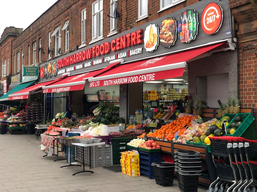 Exterior view of South Harrow Food Centre in Kingston upon Thames, featuring a red awning with white text and colorful signage indicating various food options. The storefront displays a variety of fresh produce including watermelons, oranges, apples, and other fruits arranged in green crates and baskets on the sidewalk. To the left, a small stand offers additional fruits and vegetables, with some items covered by a patterned cloth. The area is well-lit with natural daylight, showcasing clean, organized displays and a tidy pavement. The building is made of brick with large windows above the storefront, and the scene exudes a vibrant, bustling market atmosphere. The image is associated with the cleaning services provided by Cleaners Kingston upon Thames, highlighting the importance of maintaining cleanliness in commercial market environments.