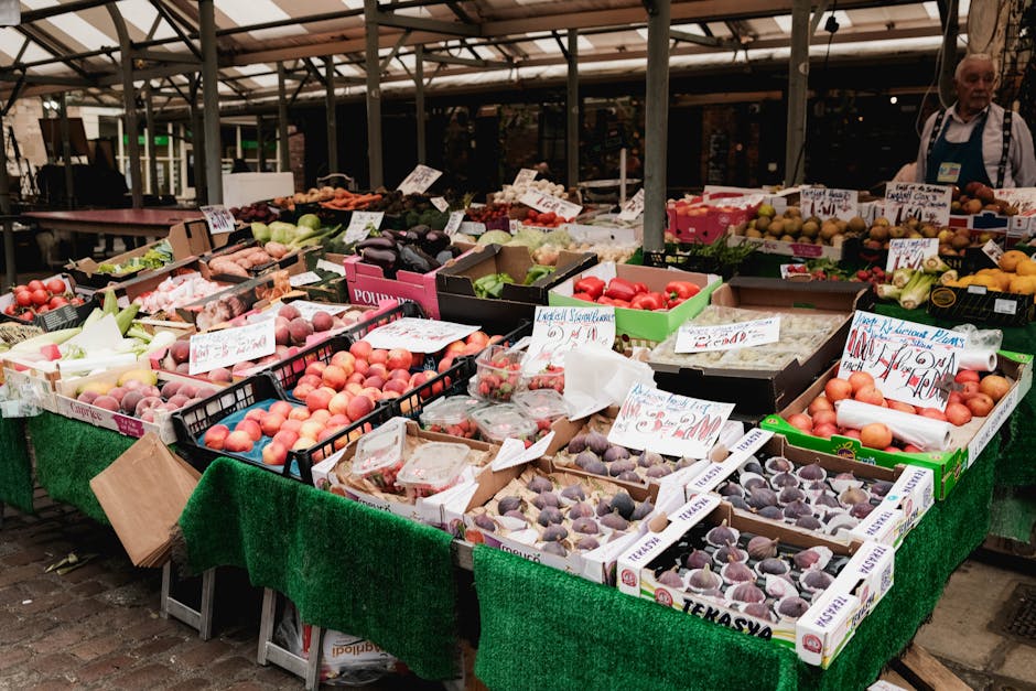 A lively outdoor market stall with multiple open cardboard boxes and crates filled with fresh fruits such as peaches, plums, and apples on a table covered with a green fabric tablecloth. The fruits are neatly arranged, some in plastic containers, with handwritten price and product labels visible. Behind the stall, there is a glass and metal structure providing shade, and a vendor wearing a white apron and striped shirt is standing to the right, overseeing the display. The scene is well-lit with natural sunlight, emphasizing the vibrant colors and cleanliness of the fruit display, reflecting good surface management and hygiene in the setting. This image is related to the cleaning and maintenance services provided by Cleaners Kingston upon Thames, as part of their cleaning category, ensuring hygienic environments for retail and market settings.